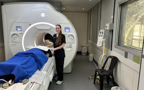 A woman in black scrubs stands next to an MRI machine with a person lying on the machine draped with a blue blanket.