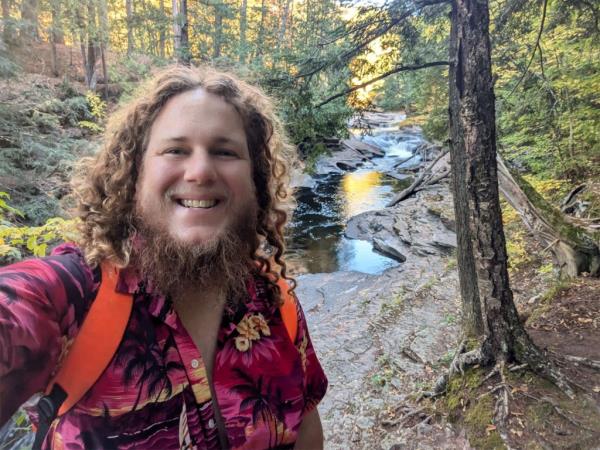 Man with long curly hair and a pink shirt, Jordan Hansen, supporting Phil Anderson for U.S. Senate in Wisconsin, standing in front of a river