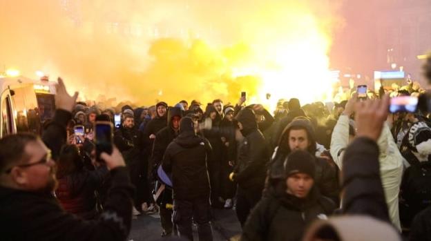 A group of men, some holding cellphones, demo<em></em>nstrate and light flares.