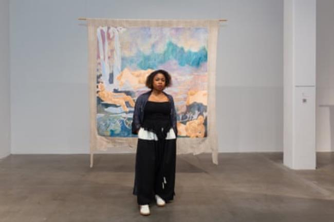 A woman stands in front of a painting that is suspended from the ceiling.